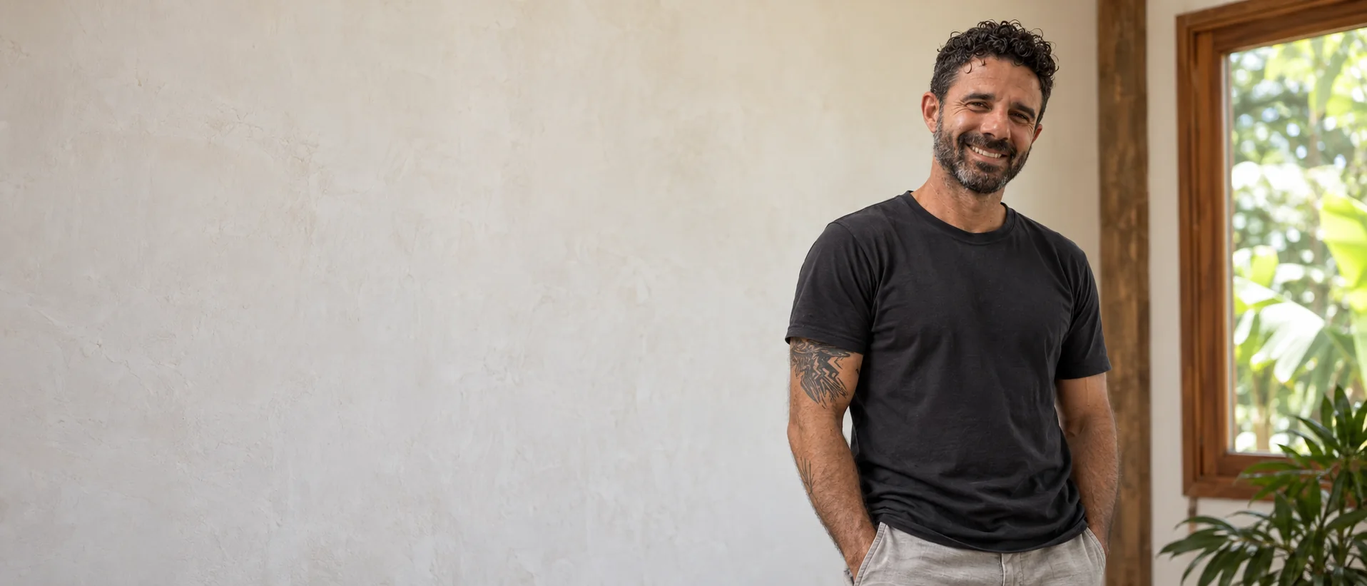 Bruno de pé, sorrindo junto à janela, de camiseta preta e com suas tatuagens nos braços
